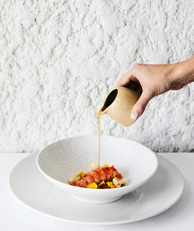 Hand holding jug of sauce being poured onto fine dining marron seafood dish at qualia Pebble Beach restaurant