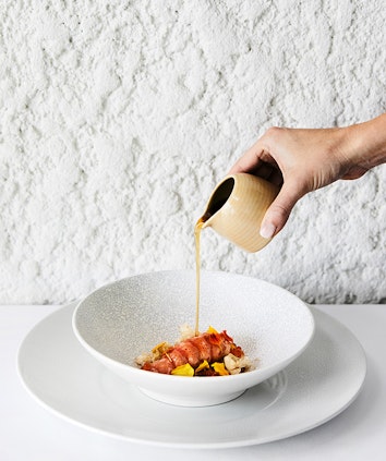 Hand holding jug of sauce being poured onto fine dining marron seafood dish at qualia Pebble Beach restaurant