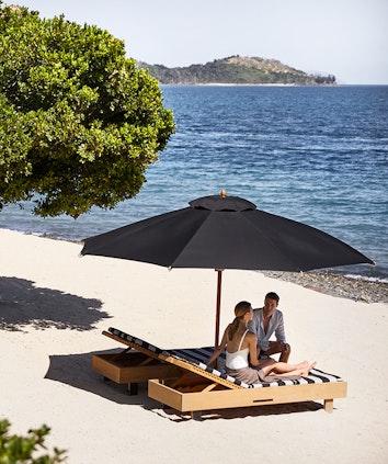 qualia Pebble Beach with man and woman sitting on striped beach lounges underneath umbrella 