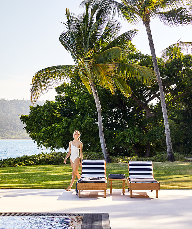 Woman in white swimsuit walking past two striped sun lounges by the Pebble Beach pool at qualia resort