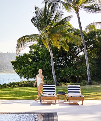 Woman in white swimsuit walking past two striped sun lounges by the Pebble Beach pool at qualia resort