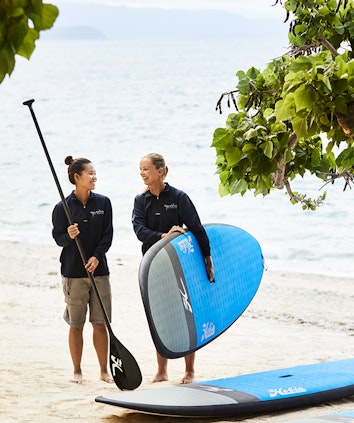 Two women part of the qualia beach sports team holding stand up paddle board equipment on the beach