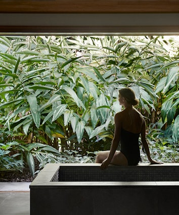 Woman sitting on edge of bathtub in spa qualia treatment room looking at lush foliage outside window