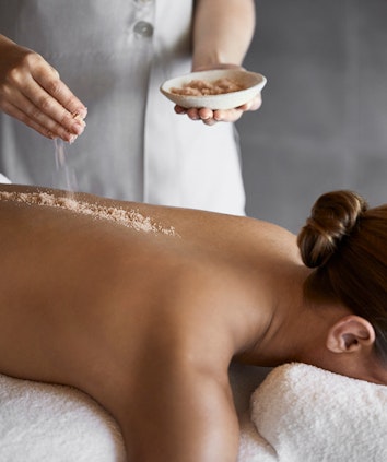 Salt scrub being sprinkled onto woman's back in spa qualia treatment room by spa therapist