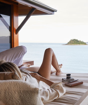 Couple laying next to camera and notebook in a day bed looking out at Whitsundays views from qualia resort pavilion