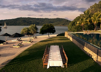 Aerial view of long table with white cloth in the grass and qualia infinity pool to the right and Pebble Beach to the left 
