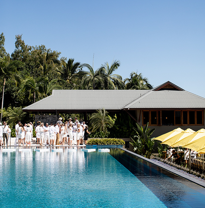View of group people dressed in white and large lunch table under yellow umbrellas from across qualia inifinity pool 