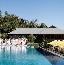 View of group people dressed in white and large lunch table under yellow umbrellas from across qualia inifinity pool 