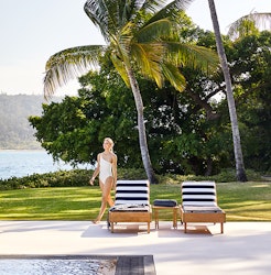 Woman in white swimsuit walking past two striped sun lounges by the Pebble Beach pool at qualia resort