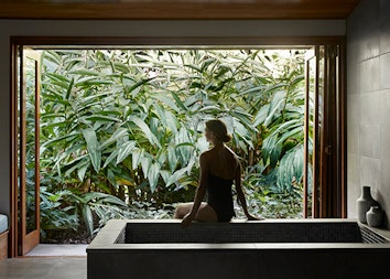 Spa qualia treatment room with woman sitting on bath edge looking out the window to views of lush foliage