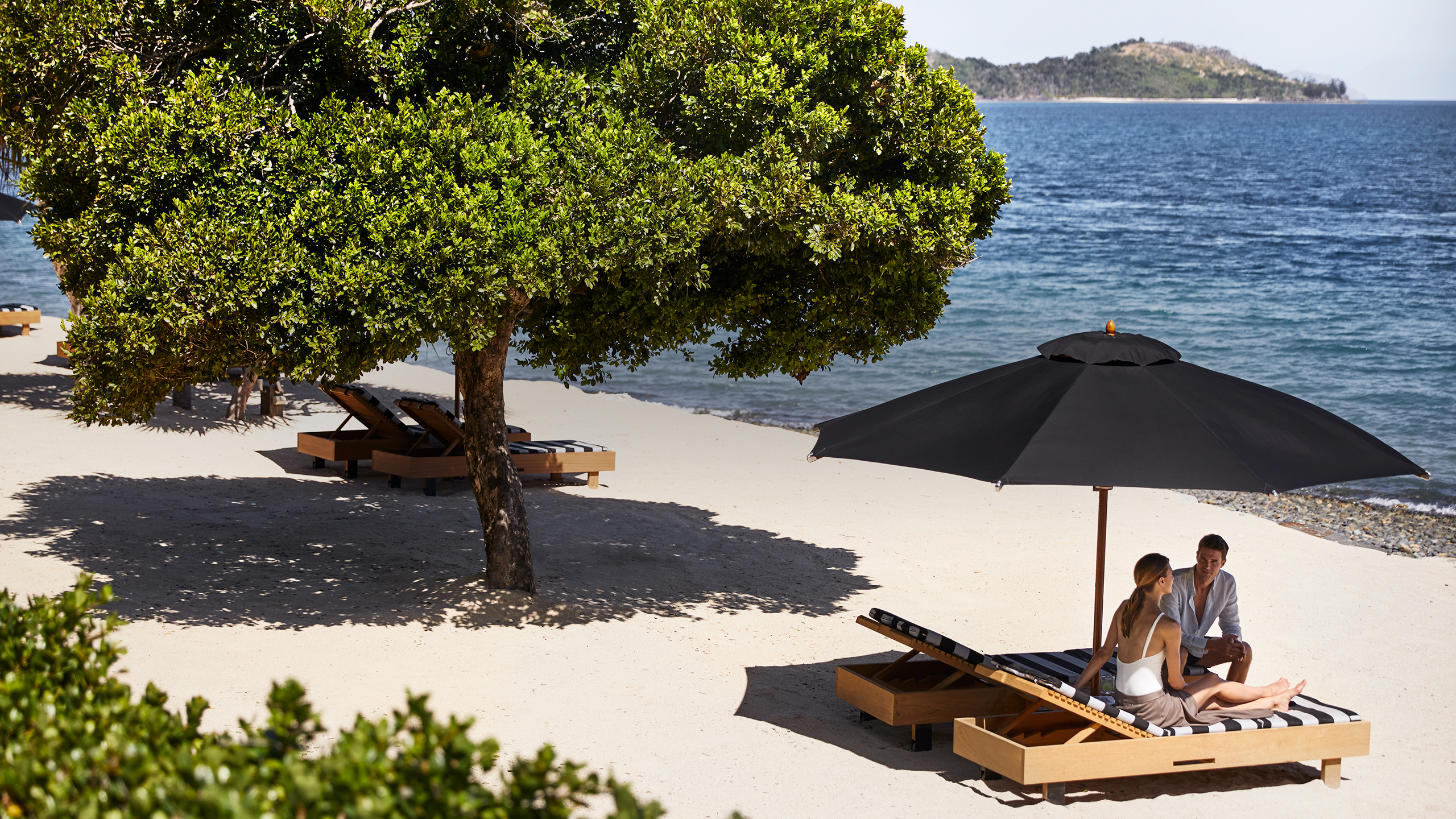 qualia Pebble Beach with man and woman sitting on striped beach lounges underneath umbrella