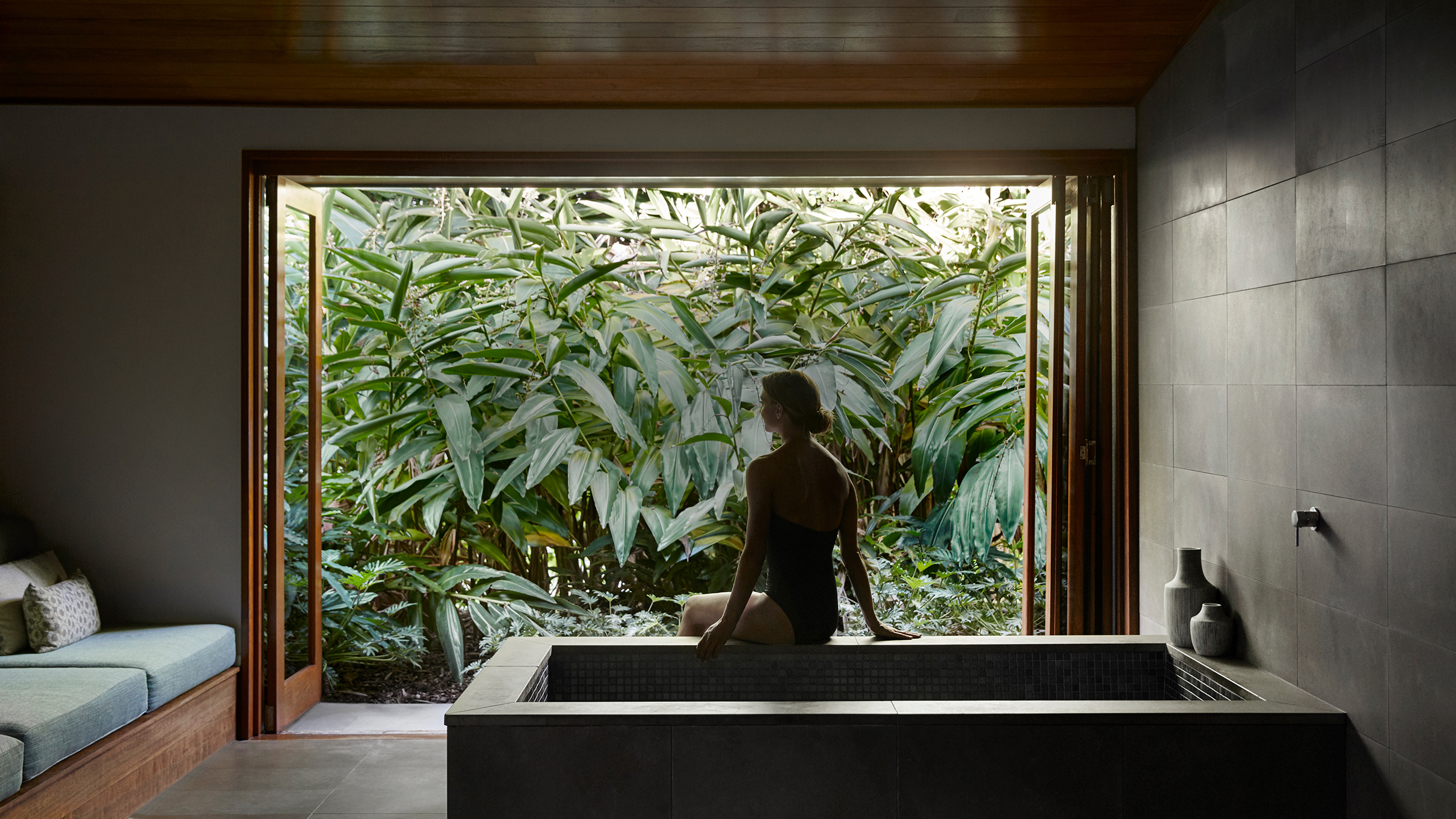 Spa qualia treatment room with woman sitting on bath edge looking out the window to views of lush foliage