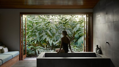 Spa qualia treatment room with woman sitting on bath edge looking out the window to views of lush foliage